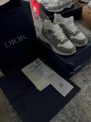 Dior b25 gråvita sneakers - Snygga Dior b25 sneakers i grått och vitt med klassiskt Dior Oblique-mönster på sidan. Ovandel i mocka och mesh, vita skosnören och robust sula. Perfekta för dig som vill sticka ut med exklusiva och trendiga skor från Dior.