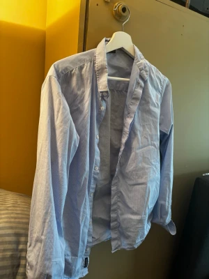 Blåvit randig skjorta med knappar - Snygg blåvit randig skjorta med klassisk krage och knäppning framtill. Skjortan har långa ärmar och är tillverkad i ett lätt bomullstyg, perfekt för en stilren look. Passar dig som gillar en enkel men fräsch stil.