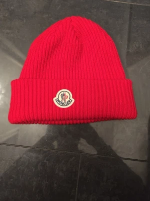 Röd Moncler mössa i ribbstickad ull - Snygg röd ribbstickad mössa från Moncler med uppvikt kant och klassisk Moncler-logga framtill. Tillverkad i mjuk ull som håller dig varm och ger en trendig look. Perfekt accessoar för kyliga dagar.