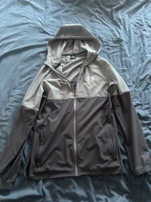 Grå vindjacka från Under Armour - Snygg tvåfärgad vindjacka från Under Armour i ljusgrått och mörkgrått. Jackan har huva, dragkedja framtill och långa ärmar. Perfekt för blåsiga dagar och sportiga outfits. Under Armour-logga på bröstet och diskreta detaljer vid ärmslut.
