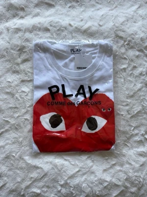 Vit Comme des Garçons Play t-shirt - Vit t-shirt från Comme des Garçons Play med det ikoniska röda hjärtat med ögon tryckt på bröstet. Klassisk rund hals och korta ärmar. Perfekt för dig som gillar streetstyle och vill sticka ut med en cool design.