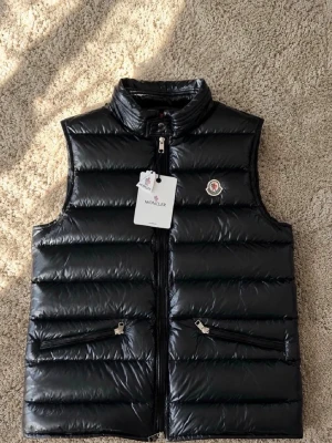 Moncler väst - Snygg svart dunväst från Moncler med glansig finish, ståkrage och två dragkedjefickor framtill. Västen har Moncler-logga på bröstet och stängs med dragkedja. Perfekt för lager-på-lager och streetstyle.