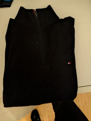 Svart halvzip från Tommy Hilfiger - Svart stickad halvzip-tröja från Tommy Hilfiger i mjuk bomull- och kashmirblandning. Klassisk design med liten logga på bröstet och ribbade detaljer med rödvita ränder i kragen. Perfekt för en stilren och bekväm look.