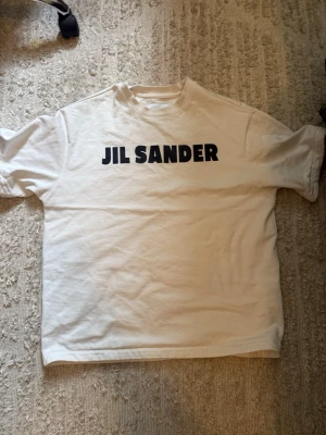 Vit Jil Sander t-shirt  - Vit t-shirt från Jil Sander med svart logotyptryck på bröstet. Klassisk rak passform och rund halsringning. Tillverkad i mjuk bomull och har korta ärmar. Perfekt för en clean och stilren look.