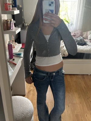 Grå croppad hoodie med dragkedja - Snygg grå croppad hoodie med dragkedja framtill och ribbade muddar. Tröjan har huva och är tillverkad i mjukt stickat material. Perfekt för en avslappnad och trendig look.( tänk på att tröjan är lite liten för mig men för en som har storlek 146/152 kanske den är till naveln)