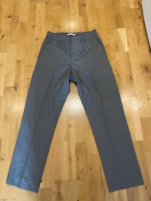 Brand Island gråmjukisbyxor med resårmidja stl S - Snygga grå mjukisbyxor med rak passform och elastisk midja. Byxorna har sidofickor och en bakficka, perfekta för en avslappnad stil. Tillverkade i ett mjukt och bekvämt material som passar för chill dagar.