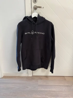 Svart hoodie från Sail Racing - Säljer en svart hoodie från Sail Racing med vit logga tryckt på bröstet. Hoodien har en stor känguruficka framtill. Bra skick.