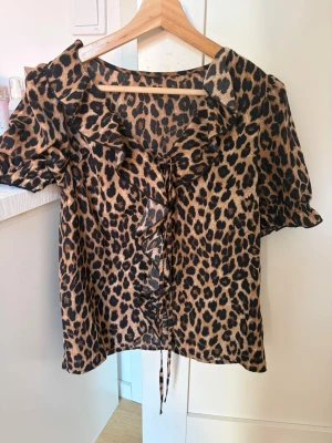 Leopardmönstrad blus med volang - Snygg blus med leopardmönster och volangdetalj framtill. Blusen har korta puffärmar och en bred krage. Materialet är glansigt och känns lyxigt, perfekt för att sticka ut. Knytband framtill för extra detalj. Xs-s