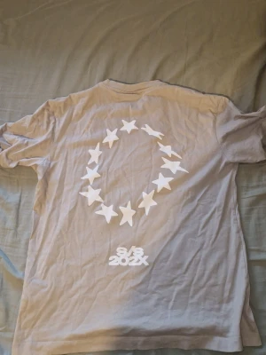Beige t-shirt med stjärntryck S/S 202X - Beige t-shirt med vita stjärnor tryckta i cirkel på ryggen och i ett rutmönster på bröstet. Tryck med texten S/S 202X både fram och bak. Klassisk passform och rund halsringning. Perfekt för en trendig och avslappnad stil.