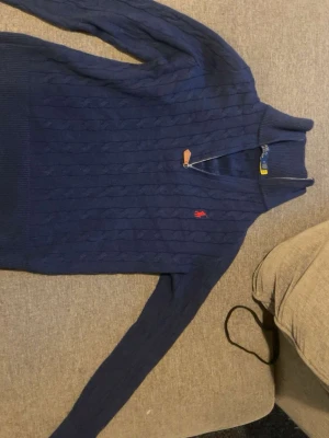 Blå kabelstickad tröja Polo Ralph Lauren - Mörkblå kabelstickad tröja från Polo Ralph Lauren med halv dragkedja och röd broderad logga på bröstet. Tröjan har hög krage och ribbade muddar. Tillverkad i mjukt material som passar perfekt för lager på lager.