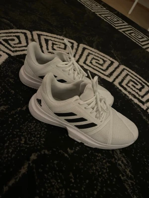 Vita Adidas sneakers med svarta ränder - Fräscha vita sneakers från Adidas med klassiska svarta ränder på sidorna. Skorna har en låg profil, snörning och ovandel i mesh för bra andningsförmåga. Perfekta för dig som gillar en clean och sportig stil.