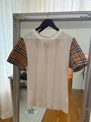 Vit t-shirt med rutiga ärmar Burberry - Vit t-shirt från Burberry med klassiska kortärmade rutiga ärmar i beige, svart och rött. Tidlös design med rund halsringning och en skön passform. Perfekt för dig som vill ha en stilren men ändå unik look. BARNSTORLEK