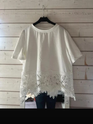 Vit topp med spetsdetaljer - Vit topp med korta ärmar och rund halsringning. Nederdelen är dekorerad med breda spetsdetaljer och broderade blommor som ger en romantisk känsla. Perfekt att styla med jeans eller kjol för en fräsch look.