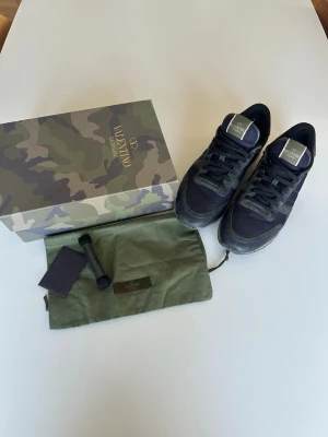 Valentino Garavani svarta sneakers - Snygga svarta sneakers från Valentino Garavani med detaljer i mocka, läder och textil. Skorna har ikoniska nitar på hälen och en robust sula. Kommer med originalkartong, dustbag och extra skosnören. Perfekta för dig som gillar exklusiv streetstyle.