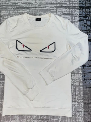 Vit Fendi sweatshirt med ögonmotiv - Cool vit sweatshirt från Fendi med unikt ögonmotiv och dragkedja framtill. Tröjan har rund hals, långa ärmar och ribbade muddar. Perfekt för dig som vill sticka ut med en stilren men edgy look.