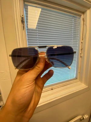 Prada blå solglasögon med metallbåge - Snygga solglasögon från Prada med blåtonade glas och silvrig metallbåge. Skalmarna är svarta med röda Prada-detaljer och vita ändar. Klassisk fyrkantig form som ger en cool vibe och passar till många stilar. Obs har lite repor på glaset där med priset 