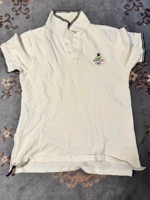 Vit pikétröja med OS-brodyr - Vit pikétröja med svart kant på krage och ärmslut. Broderad logga från Olympiska spelen i Sydney 2000 på bröstet. Klassisk pikékrage med knappar framtill och korta ärmar. Perfekt för dig som gillar sportig stil.