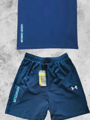 Blått träningsset från Under Armour - Snyggt träningsset med t-shirt och shorts i mörkblått från Under Armour. Båda plaggen har logga och text i turkos och vitt. Shortsen har resår i midjan och färgglad logotyp på benet. Perfekt för gym eller löpning. Pris kan diskuteras vid snabb affär