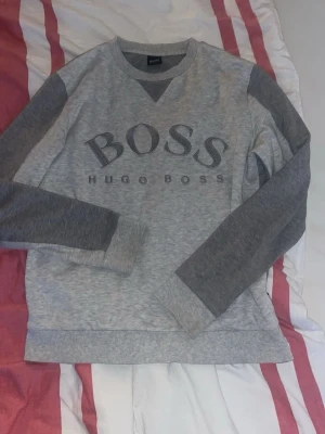 Grå sweatshirt från Hugo Boss - Snygg grå sweatshirt från Hugo Boss med mörkare grå ärmar och ribbade muddar. Stor BOSS-logga framtill och klassisk rund halsringning med V-detalj. Perfekt för en avslappnad och trendig stil.