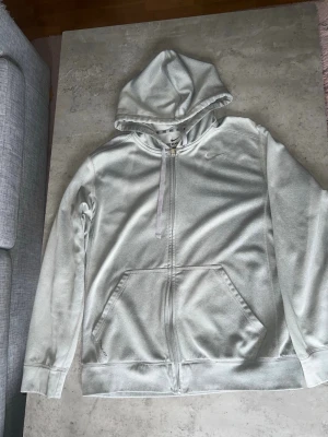 Ljusgrå zip-hoodie från Nike - Ljusgrå hoodie från Nike med dragkedja framtill och klassisk huva. Tröjan har två stora fickor på magen och en diskret broderad Nike-logga på bröstet. Tillverkad i mjukt och bekvämt material, perfekt för en avslappnad stil.