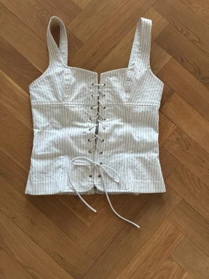 Vit randig korsett-topp med snörning - Trendig vit korsett-topp med tunna ränder och snörning framtill. Breda axelband och figurnära passform. Perfekt att styla med jeans eller kjol för en cool look.