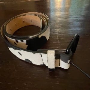 Svartvitt bälte i skinn med ko-mönster - Snyggt bälte i svartvitt ko-mönster med svart spänne. Bältet är gjort i skinn och har en naturlig insida. Perfekt accessoar för att lyfta din outfit med en cool touch!!