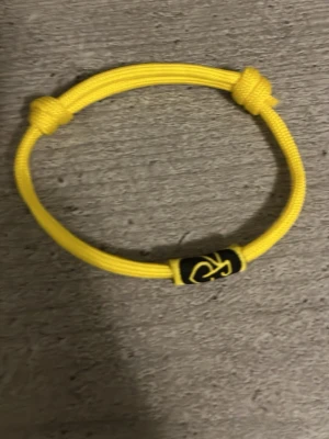 Gult justerbart armband i textil - Snyggt gult armband i slitstark textil med justerbara knutar och en svart detalj med gul logga. Perfekt för dig som gillar enkel och sportig stil. Passar både ensam eller tillsammans med andra armband för en personlig look.