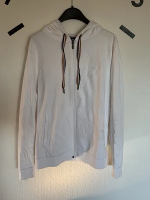 Vit hoodie med dragkedja från BOSS - Snygg vit hoodie från BOSS med dragkedja framtill och två fickor. Huva med justerbara snören i svart och brunt. Diskret BOSS-logga broderad på bröstet. Tillverkad i mjukt och lätt material, perfekt för en clean och stilren look.