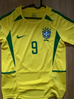 Brasilien fotbollströja - Gul Brasilien fotbollströja från Nike med gröna detaljer och nummer 9 på bröstet. Tröjan har rund hals, broderat CBF-emblem och är tillverkad i lätt och ventilerande material som passar perfekt för fotbollsträning.