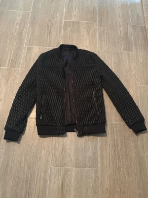 Svart quiltad bomberjacka Zara Man - Snygg svart bomberjacka från Zara Man med quiltat mönster och ribbade muddar vid ärmslut och midja. Jackan har dragkedja framtill och två fickor med dragkedja. Perfekt för dig som vill ha en stilren och trendig look.