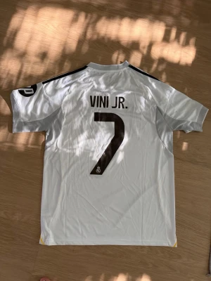 Real Madrid matchtröja Vini Jr. 7 - Snygg vit Real Madrid matchtröja från Adidas med Vini Jr. och nummer 7 på ryggen. Tröjan har svarta detaljer, gula linjer längs sidorna och klubbmärke samt sponsortryck på bröstet. Korta ärmar och sportig passform, perfekt för fotboll eller träning.