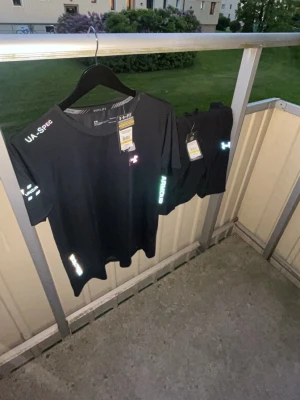 Svart träningsset från Under Armour - Snyggt svart träningsset från Under Armour med t-shirt och shorts. Setet har reflekterande detaljer och loggor på både tröja och shorts. Materialet är lätt och andas, perfekt för intensiva träningspass. Både t-shirt och shorts har märkets tryck på ärm och ben.