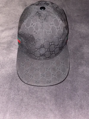 Svart keps från Gucci med monogram - Svart keps från Gucci med klassiskt GG-monogram över hela kepsen. På sidan finns en grön och röd rand som ger en ikonisk touch. Kepsen har böjd skärm och är tillverkad i tygmaterial. Perfekt för dig som gillar exklusiva accessoarer.
