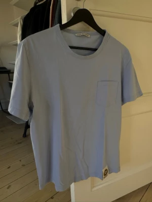 Ljusblå t-shirt från Tiger of Sweden - Snygg ljusblå t-shirt från Tiger of Sweden med rund halsringning och bröstficka. Tillverkad i mjuk bomull, perfekt för en clean och stilren look. Klassisk passform och diskret logga nertill.