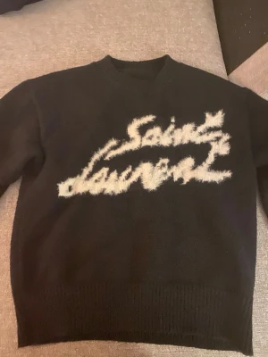 Svart stickad Saint Laurent-tröja - Snygg svart stickad tröja från Saint Laurent med vit, fluffig logotyp i graffiti-stil över bröstet. Tröjan har rund halsringning och ribbade muddar vid ärmslut och nederkant. Perfekt för dig som gillar streetstyle och exklusiva märken.