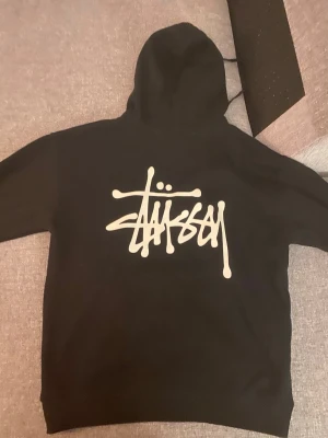 Svart Stussy hoodie - Svart hoodie från Stüssy med stor vit logga på ryggen och mindre logga på bröstet. Klassisk känguruficka framtill och huva. Perfekt för en streetwear-look. Mjuk bomullsblandning som är skön att bära.