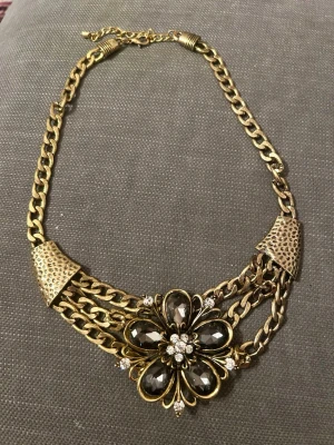 Guldfärgat statement-halsband med blomma - Snyggt statement-halsband i guldfärgad metall med kraftiga kedjor och en stor blomma i mitten. Blomman är dekorerad med svarta och klara stenar som ger en lyxig känsla. Perfekt för att lyfta vilken outfit som helst med en cool och trendig touch.