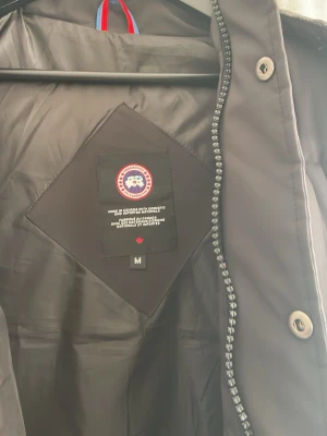 Svart dunjacka från Canada Goose - Säljer en svart dunjacka från Canada Goose med klassisk logga på insidan. Jackan har dragkedja och knappar framtill samt detaljer i hög kvalitet. Perfekt för kalla vinterdagar och riktigt varm tack vare dunfyllningen.