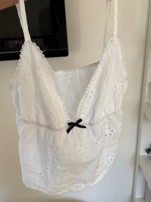 Vit broderad spetslinne med rosett - Söt vit topp med broderade blommor och tunna axelband. Djup V-ringning med spetskant och liten svart rosett framtill. Lätt och luftig bomullskänsla, perfekt för en romantisk look.