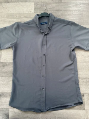 Grå kortärmad skjorta från New Look - Stilren grå skjorta från New Look med korta ärmar och klassisk krage. Skjortan har knäppning framtill och är tillverkad i ett mjukt material som känns bekvämt mot huden. Perfekt för en clean och enkel look.