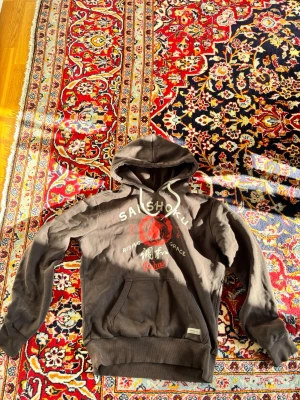 Brun hoodie från superdry  - Cool brun hoodie från Superdry med stor huva och vitt snöre. Framsidan har ett rött och vitt tryck med text och japanska tecken. Mjuk insida och känguruficka framtill. Perfekt för en avslappnad streetstyle-look.