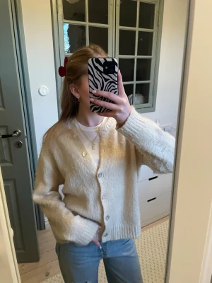 Beige stickad kofta med knappar - En beige stickad kofta med v-ringning och pärlemorknappar. Koftan har långa ärmar och ribbade muddar vid ärmslut och nederkant. Materialet ser ut att vara mjukt och lite fluffigt, perfekt för lager-på-lager och chill stil.