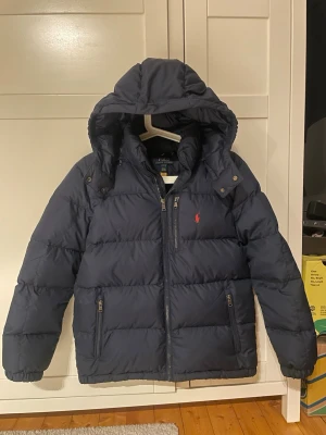 Mörkblå dunjacka med huva - Storlek L i barn 14-15 år. Skriv vid minsta fråga. Snygg mörkblå dunjacka med huva och breda kanaler. Jackan är vadderad och har en klassisk pufferstil som håller dig varm under kalla dagar. Perfekt för vintern och har en enkel, stilren design utan synliga loggor.