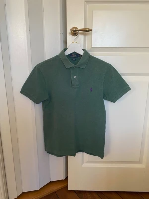 Grön pikétröja från Ralph Lauren - Klassisk grön pikétröja från Ralph Lauren med korta ärmar och krage. Tröjan har två knappar vid halsen och är mycket skön att bära.