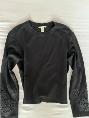 Svart långärmad topp med spetsdetaljer - Svart långärmad topp från H&M med rund halsringning och snygga spetsdetaljer på ärmarna. Tillverkad i mjuk bomull som känns skön mot huden. Perfekt för dig som gillar stilrena plagg med en twist.