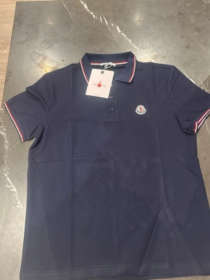 Marinblå Moncler pikétröja - Snygg marinblå pikétröja från Moncler med klassisk krage och knäppning framtill. Tröjan har röda och vita detaljer på krage och ärmslut samt Moncler-logga på bröstet. Perfekt för en stilren look.