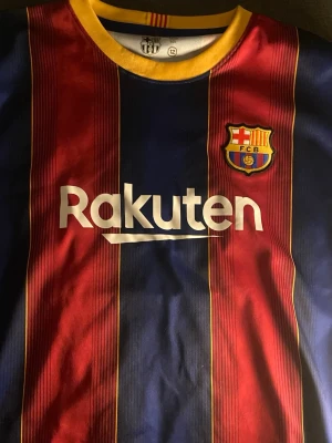 FC Barcelona Messi matchtröja - Säljer en officiell FC Barcelona matchtröja med Messi 10 på ryggen. Tröjan har klassiska blå och röda vertikala ränder, gula detaljer vid kragen och tryck från Rakuten och UNICEF. Perfekt för dig som älskar fotboll och vill hylla Messi.