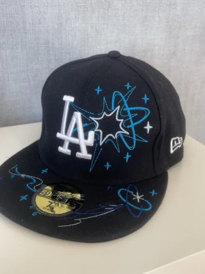 Svart LA-keps från New Era 59Fifty 7 1/4 57.8cm - Snygg svart keps från New Era 59Fifty med LA-logga framtill. Kepsen har broderade blå och vita rymdmotiv och stjärnor både fram och bak. Platt skärm och klassisk passform. Perfekt för dig som gillar streetwear och sportig stil. 