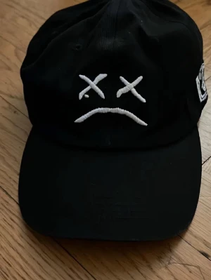 Svart keps med broderad smiley - Säljer en svart keps med böjd skärm och broderad vit smiley med korsade ögon och nedåtvänd mun på framsidan. Kepsen har justerbar rem bak och är tillverkad i bomull. Perfekt för en avslappnad streetwear-look.
