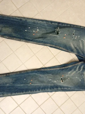 Dsquared2 blå slitna jeans - Säljer ett par blå jeans från Dsquared2 med slitningar, hål och färgstänk för en riktigt edgy look. Jeansen har raka ben och klassiska bakfickor med detaljerade sömmar. Perfekta för dig som gillar streetstyle och vill sticka ut. Passar 165-175